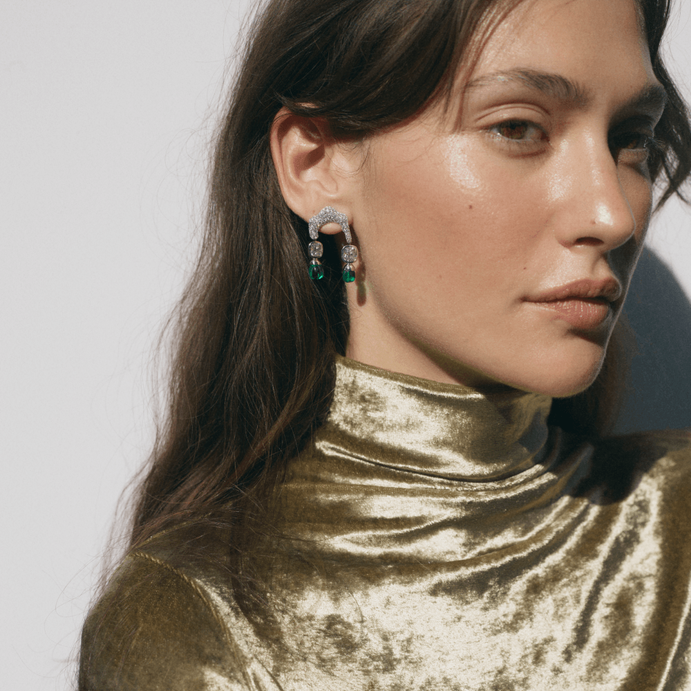 A close-up of a woman wearing a textured metallic gold turtleneck top and a dangling hoop earring with clear stones and green gemstone accents, set against a bright neutral background.