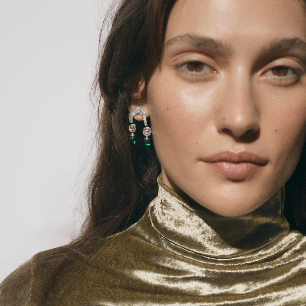 A close-up portrait of a woman wearing a pair of silver-toned statement earrings with green gemstone drops, paired with a textured olive green velvet turtleneck top against a plain light background.